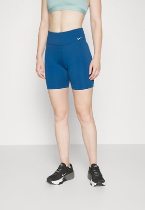 Frau trägt blaue, hoch taillierte Nike-Biker-Shorts, schwarz-weiße Nike-Sneaker und einen hellgrünen Sport-BH, steht vor einer weißen Wand.