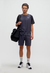 Νεαρός άνδρας με μαύρα και γκρι αθλητικά ρούχα κρατώντας μια μαύρη τσάντα γυμναστηρίου, στέκεται μπροστά σε απλό φωτεινό φόντο.