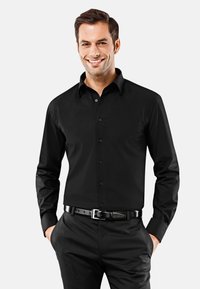 Chemise noire à manches longues, boutonnée, en tissu lisse, avec col pointu et fermeture à boutons sur le devant, portée avec un pantalon sombre et une ceinture.