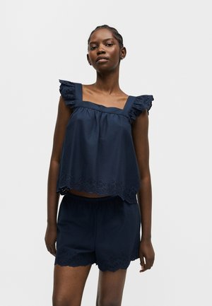 Žena v tmavomodrom tielku bez rukávov a krátkych nohaviciach s vlnitými krajkami s očkami, stojaca na jednoduchom svetlom pozadí.