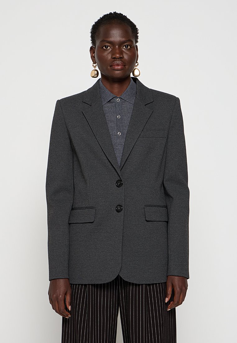 Blazer grigio in misto lana strutturato, monopetto con due bottoni, revers a punta e due tasche frontali. Abbinato a una camicia con colletto.