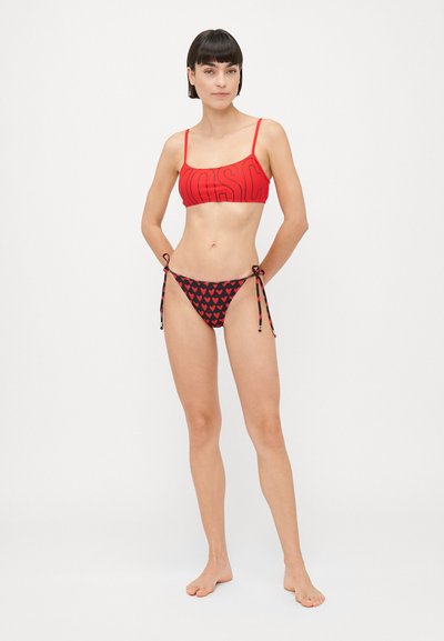 Sieviete stāv basām kājām sarkanā īsā bikini augšdaļā ar daļēju logo un melnās bikini apakšdaļā ar sarkanu sirdīšu rakstu, kas piesietas sānos.