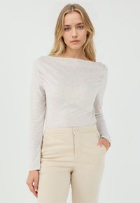 Langermige, off-the-shoulder top in lichtbeige stof met een zachte textuur, gecombineerd met beige hooggesneden broeken met frontzakken.