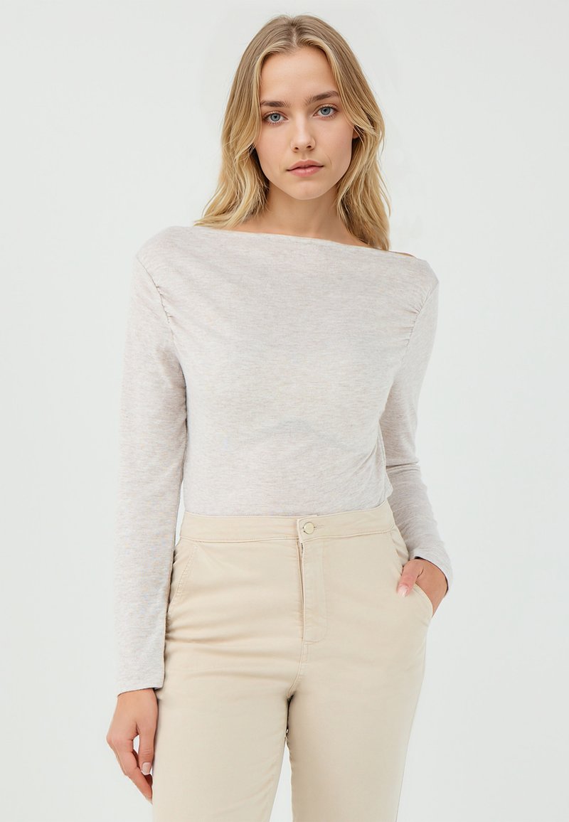 Langermige, off-the-shoulder top in lichtbeige stof met een zachte textuur, gecombineerd met beige hooggesneden broeken met frontzakken.