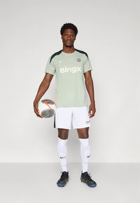 Mees jalgpallur Chelsea treeningvormis, hoides jalgpalli, kandes valgeid pükse, sokke ja musti krampe, seistes ühevärvilise tausta ees.