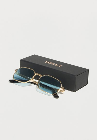 Γυαλιά ηλίου Versace με χρυσό πλαίσιο και φακούς με μπλε απόχρωση, τοποθετημένα μπροστά σε μια μαύρη θήκη με το λογότυπο Versace πάνω σε λευκή επιφάνεια.