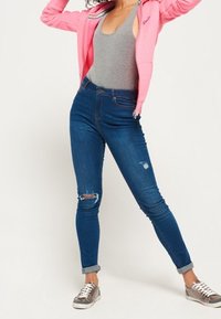 Sudadera con cremallera de color rosa claro, camiseta sin mangas gris y jeans ajustados de color azul oscuro con la rodilla rasgada, combinados con zapatillas metálicas plateadas.