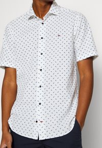 Camicetta bianca a maniche corte con motivo geometrico blu navy, colletto button-down e delicato dettaglio del logo sul petto. Realizzata in tessuto leggero.