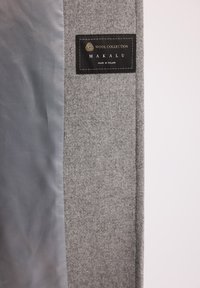 Czarna etykieta "Wool Collection Makalu Made in Poland" na szarej wełnianej tkaninie z gładkim szarym podszyciem wewnątrz płaszcza lub kurtki.