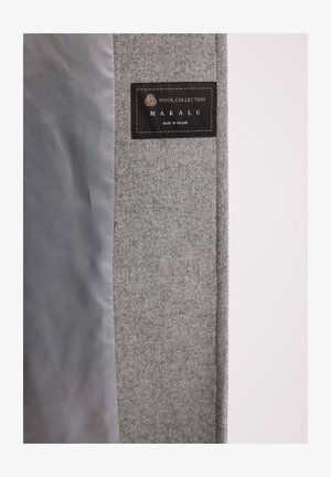 Czarna etykieta "Wool Collection Makalu Made in Poland" na szarej wełnianej tkaninie z gładkim szarym podszyciem wewnątrz płaszcza lub kurtki.