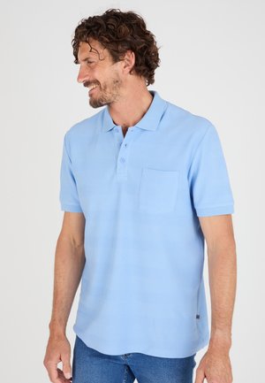 Hombre con pelo rizado que lleva una camiseta tipo polo de manga corta azul clara y vaqueros azules, sonriendo y mirando hacia su izquierda.