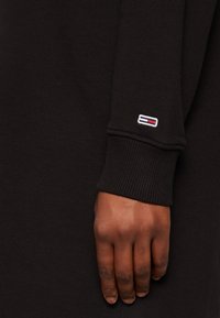 Czarny materiał sweatshirtu z żebrowanymi mankietami. Wyraźne logo w białym, czerwonym i niebieskim kolorze wyszyte na rękawie. Zbliżenie na dłoń na tle tkaniny.