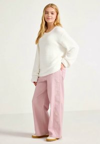Pull blanc en maille duveteuse associé à un pantalon ample rose clair avec une broderie latérale et une coupe large. Chaussures à semelles plates blanches.