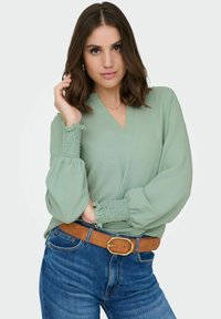 Blouse vert clair avec un col en V, manches longues smockées et tissu texturé, assortie à un jean bleu et une ceinture marron avec une boucle en or.