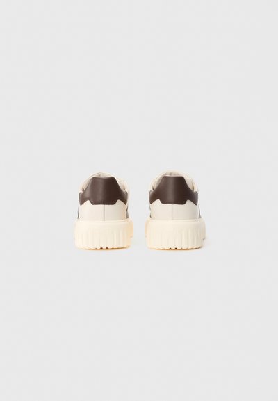 Un paio di sneakers crema con suole spesse e rigate e tappi del tallone marrone scuro, mostrati da dietro su uno sfondo chiaro e semplice.