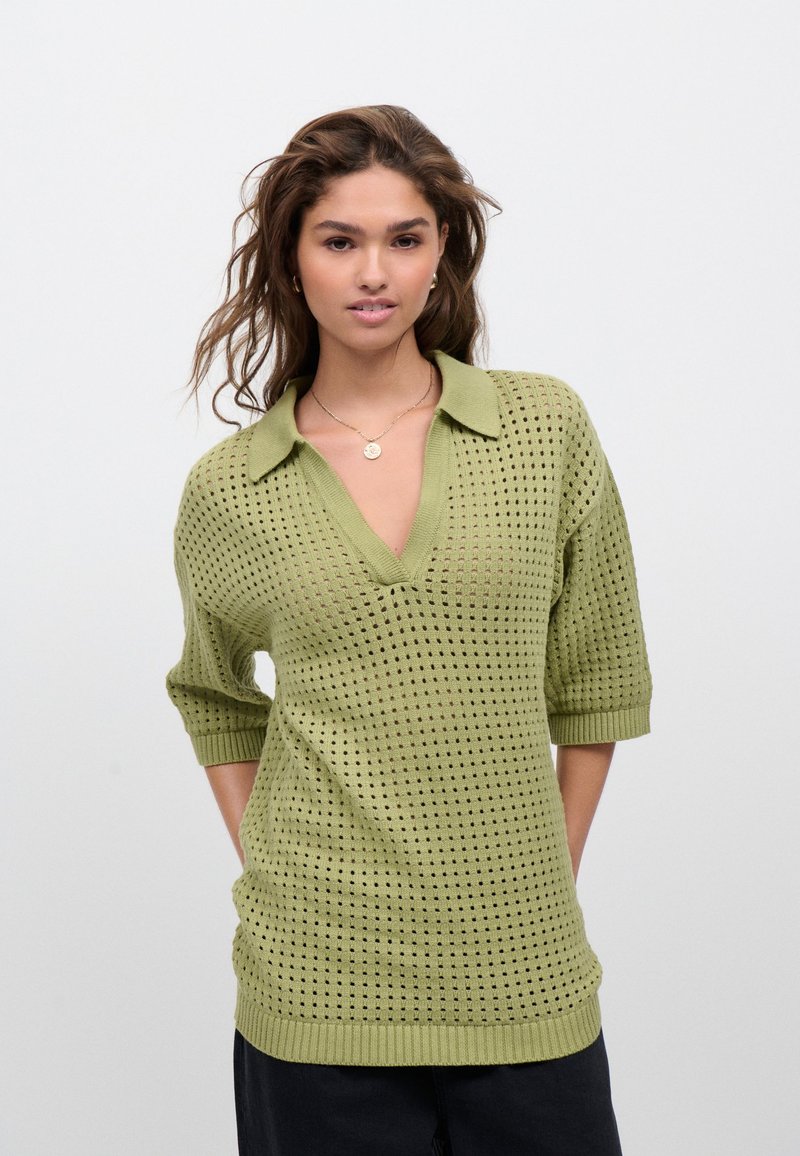 Maglione corto a maniche corte di colore verde chiaro, a maglia, con scollo a V e motivo traforato. Dettagli a coste sul colletto e sull'orlo, vestibilità rilassata.