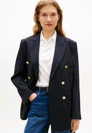 Dunkel marineblaues, doppelt geknöpftes Sakko mit goldenen Knöpfen, mit strukturierten Schultern und glattem Stoff, kombiniert mit einem weißen Polo und Jeans.