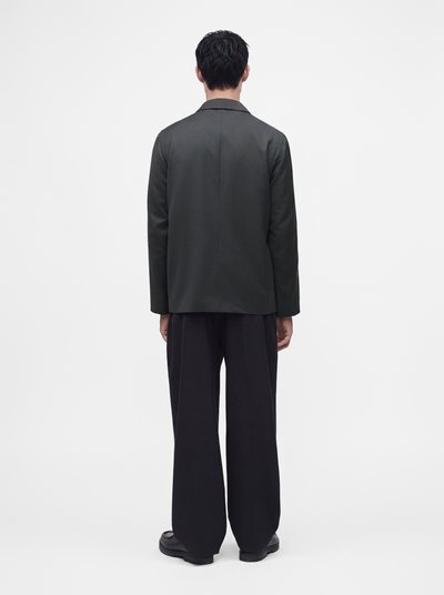 Un homme est debout, de dos, portant un blazer vert foncé, un pantalon noir ample et des chaussures noires sur un fond clair uni.