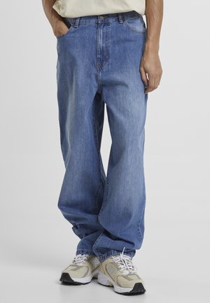 Personne portant un jean bleu ample avec des baskets beige et blanches, debout avec une main dans la poche devant un fond uni.