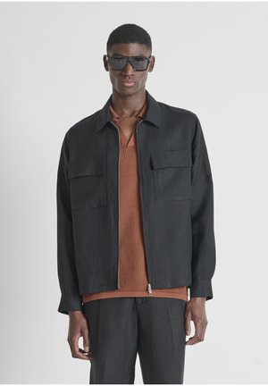 Hombre vistiendo una chaqueta negra de cremallera extragrande y pantalones a juego, camisa marrón con textura debajo y grandes gafas de sol negras, de pie frente a un fondo blanco.