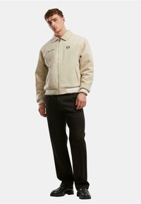 Bežinė fliso striukė su užtrauktuku priekyje, per dideliu apykakle ir siuvinėtu logotipu. Suderinta su juodomis kelnėmis ir juodais batais.