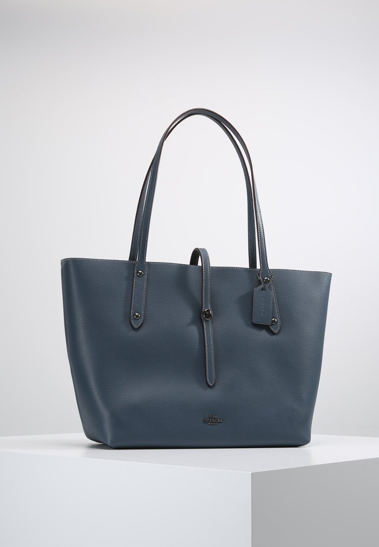 Bolso tote de piel azul con dos asas largas, superficie texturizada, acentos de hardware plateado y una pequeña etiqueta adjunta. Diseño espacioso y estructurado.