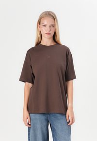 T-shirt oversized castanho em algodão macio, com mangas curtas, decote redondo e detalhe discreto do logótipo na frente. Combinado com jeans de perna larga azuis.