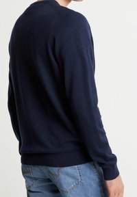 Mörkblå, stickad tröja med rund halsringning och ribbat nederkant, sedd bakifrån, i kombination med ljusblå denimjeans.