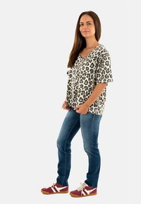Top de manga corta con estampado de leopardo en beige y marrón, combinado con jeans azules y zapatillas rojas, de pie sobre un fondo blanco.