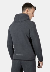 Grå hettegenser med raglanermer, som har en subtil tekstur, logo trykt på baksiden, og en tettsittende design rundt livet.