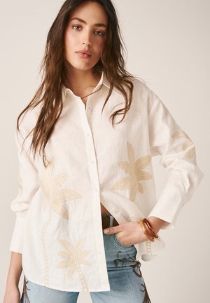 Mujer con una camisa blanca de botones, suelta, con estampados de palmeras beige y vaqueros azul claro bordados con diseños marrón oscuro.