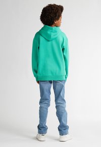 Felpa verde con cappuccio foderato, abbinata a jeans azzurri e sneakers bianche. Il tessuto sembra morbido e informale.