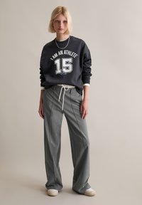 Czarna bluza z białym napisem "JESTEM SPORTOWCEM 15", w połączeniu z szarymi szerokimi spodniami i białymi trampkami. Gładka tekstura, luźny krój.