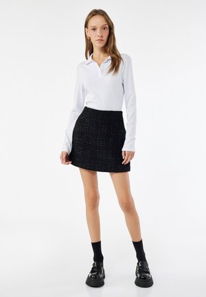 Witte lange mouw polo shirt gecombineerd met een zwart gestructureerde mini rok. Model draagt zwarte sokken en glanzende plateau schoenen voor een complete look.