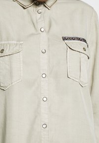 Chemise beige boutonnée avec deux poches poitrine, dont une avec un rabat et une bordure décorative le long du bord supérieur.