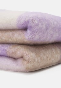 Couverture en tissu duveteux pliée, beige et violet clair, à texture douce sur une surface blanche.