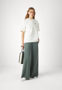 Felpa bianca a maniche corte con un piccolo logo verde, abbinata a pantaloni larghi verdi. Gli accessori includono una borsa nera e crema.