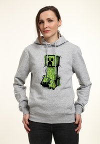 Sudadera gris con capucha que presenta un gráfico de un personaje verde, de forma cuadrada, con contornos negros y patrones intrincados en su superficie, mostrado en la parte frontal.