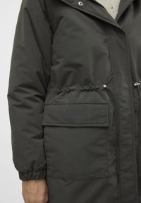 Długa, ciemnozielona kurtka puchowa z talią ściąganą, elastycznymi mankietami i przednią kieszenią. Posiada zapięcie na zamek oraz obszerny kaptur.