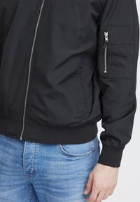 Svart bomberjakke med glidelås foran, ribbede mansjetter og en lomme på armen. Materialet virker glattere og lett, kombinert med blå denim jeans.