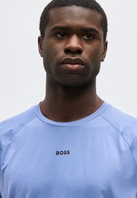 Νεαρός με κοντά σγουρά μαλλιά που φοράει ανοιχτό μπλε πουκάμισο με την επιγραφή "BOSS" σε μαύρο στο στήθος, ουδέτερη έκφραση, λευκό φόντο.