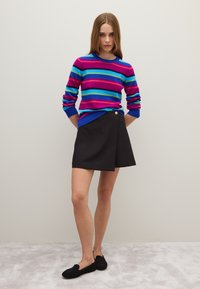 Maglione a righe multicolori in rosa, blu e verde; gonna nera avvolgente con bottone dorato; scarpe nere slip-on; sfondo neutro.