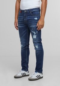 Jeans en denim bleu foncé avec des détails usés, dotés d'accents de déchirure et d'un délavage. Portés avec des baskets blanches et un débardeur blanc ajusté.