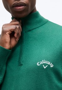 Άτομο που φοράει πράσινο πουλόβερ Callaway με φερμουάρ στο ένα τέταρτο, κρατώντας το φερμουάρ κοντά στον λαιμό, εμφανίζοντας το κάτω μέρος του προσώπου και το χέρι.