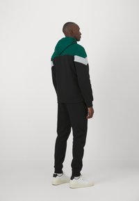 Conjunto de sudadera con capucha negra y joggers que presentan una capucha verde, acentos en bloques de color gris y verde, y puños elásticos. Combinado con zapatillas blancas.