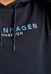 Tmavě modrá mikina s kapucí a klokaní kapsou, s nápisy v světle modré a bílé barvě, které znějí "COPENHAGEN" a "FRANDBERGH." Nastavitelné šňůrky na stažení.