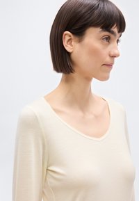 Γυναίκα με ίσιο σκούρο καστανό καρέ κούρεμα, φορώντας μπεζ μπλούζα με μακριά μανίκια, στραμμένη προς τα δεξιά μπροστά σε απλό φωτεινό φόντο.