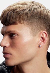 Silverfärgad studsörhänge som bärs i vänster öra, med en cirkulär design. Personen har kort, texturerat blont hår och klar hy.