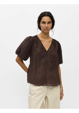 Kobieta w luźnej brązowej bluzce z dekoltem w serek i krótkich bufiastych rękawkach oraz kremowych spodniach, stojąca na jednolitym białym tle.