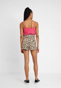 Femme aux longs cheveux tressés portant un haut rose sans manches, un short imprimé léopard et des baskets noires, debout de dos sur fond blanc.